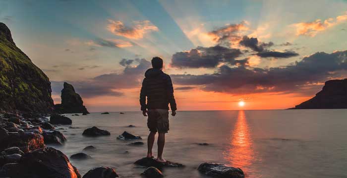 Boy standing on rocks in sea looking at sunset