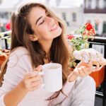 Happy young woman smiling and holding a mug of coffee and a croissant while sitting in her balcony