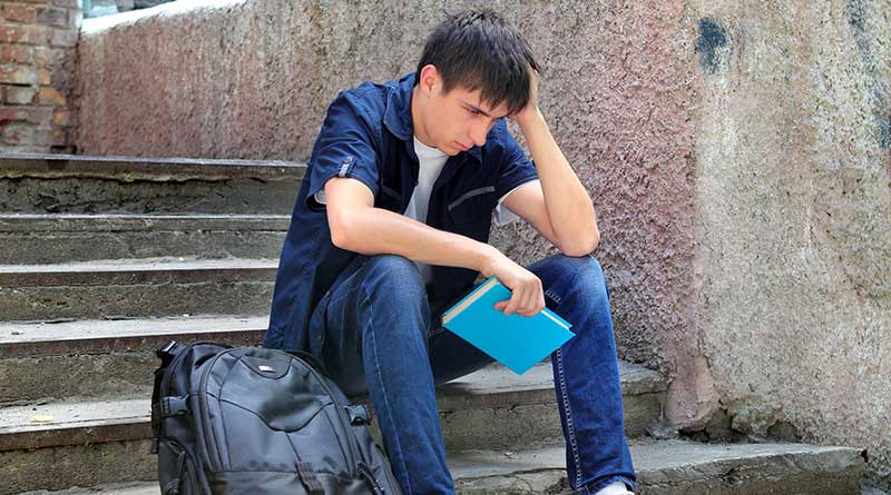 Sad student sitting on the steps of a house with his backpack