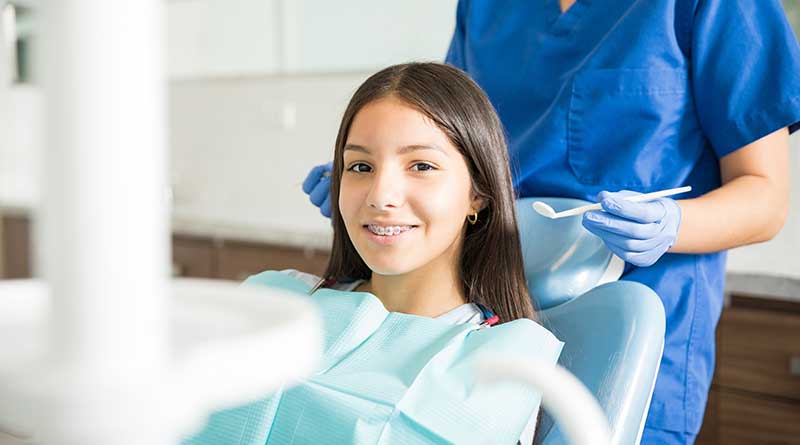 Smiling teenage girl with braces sitting at a clinic with dentist standing behind her.