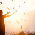 Woman standing with her arms outstretched at sunset with birds flying around