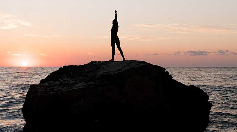 Young woman with raised arm standing on top of a rock in the ocean