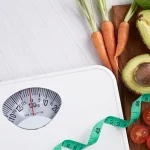 Weighing scale with measuring tape across it, and vegetables at the side of it.