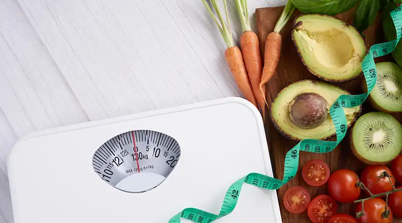 Weighing scale with measuring tape across it, and vegetables at the side of it.