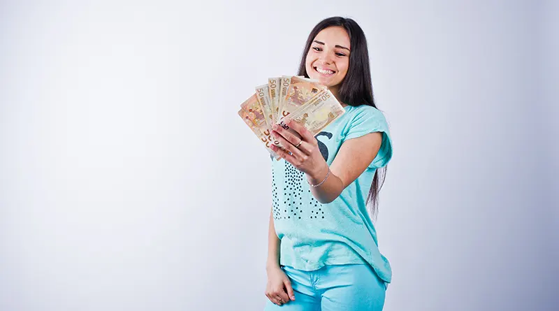 Young woman wearing a turquoise tshirt and trousers holding currency notes in her hand and smiling